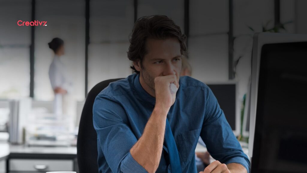 Business professional sitting at a desk, looking focused and slightly overwhelmed while working on a computer in a modern office environment.